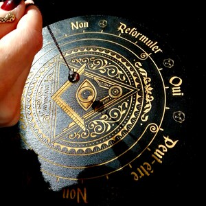 May include: A black wooden pendulum board with gold lettering and an all-seeing eye design. The board has the words "Oui", "Non", "Reformuler", and "Peut-&ecirc;tre" around the edge. A pendulum is hanging from the center of the board.