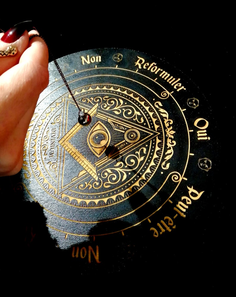 May include: A black wooden pendulum board with gold lettering and an all-seeing eye design. The board has the words "Oui", "Non", "Reformuler", and "Peut-&ecirc;tre" around the edge. A pendulum is hanging from the center of the board.