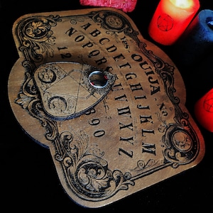 May include: A wooden Ouija board with black lettering and ornate scrollwork. The planchette is in place, and the board is surrounded by red and blue candles. A bundle of dried herbs sits nearby.