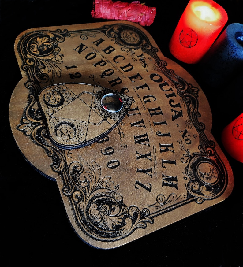 May include: A wooden Ouija board with black lettering and ornate scrollwork. The planchette is in place, and the board is surrounded by red and blue candles. A bundle of dried herbs sits nearby.