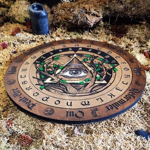 May include: A wooden Ouija board with a black and gold all-seeing eye design. The board has a black and white alphabet, numbers, and the words "Oui", "Peut-&ecirc;tre", "Non", and "Reformuler Qui".