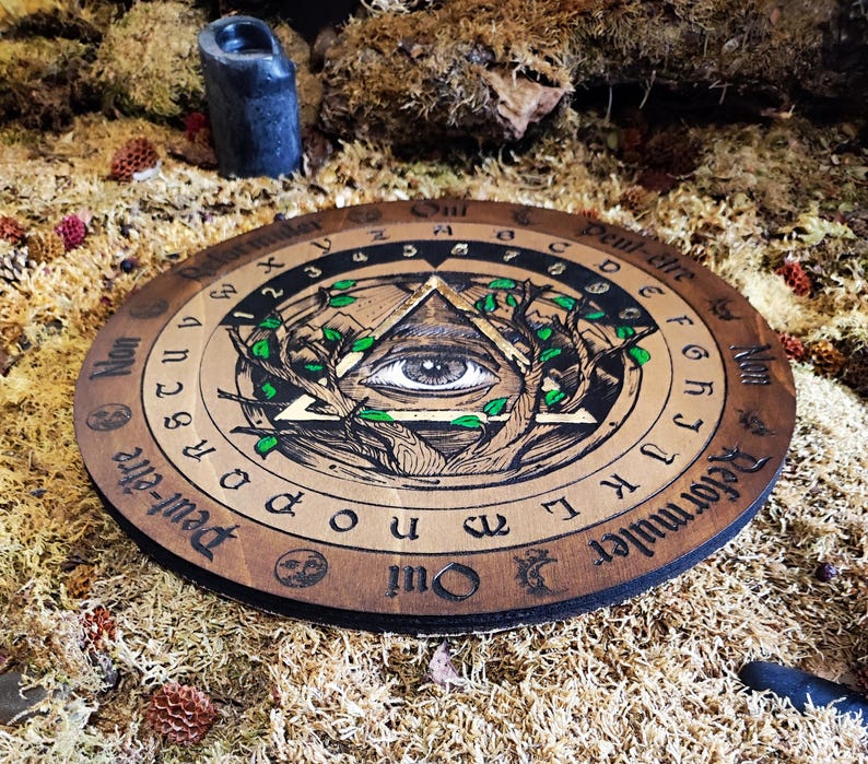 May include: A wooden Ouija board with a black and gold all-seeing eye design. The board has a black and white alphabet, numbers, and the words "Oui", "Peut-&ecirc;tre", "Non", and "Reformuler Qui".