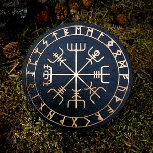 May include: A black wooden circle with a gold compass design and runes around the edge. The compass features a central star with four points, each with a different rune symbol. The runes around the edge are in a circular pattern.
