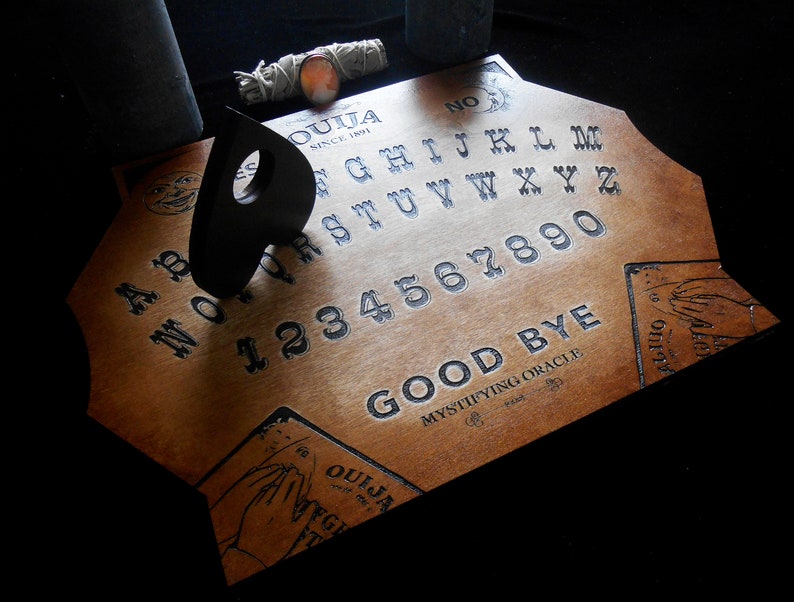 May include: A wooden Ouija board with a black planchette on top. The board has the letters of the alphabet, numbers 1-9 and 0, and the words "Good Bye" printed on it. The board is labeled "Ouija Since 1891" and "Mystifying Oracle".
