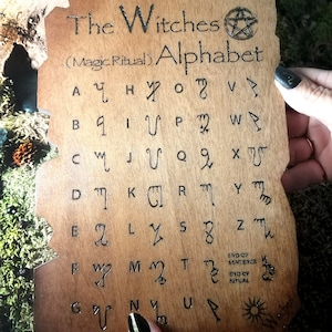 Puede incluir: Una placa de madera con el texto "The Witches (Magic Ritual) Alphabet" y un s&iacute;mbolo de pent&aacute;gono. La placa presenta una cuadr&iacute;cula de 26 letras del alfabeto, cada una con un s&iacute;mbolo &uacute;nico.