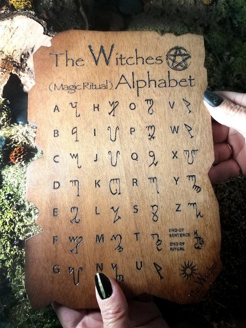 Puede incluir: Una placa de madera con el texto "The Witches (Magic Ritual) Alphabet" y un s&iacute;mbolo de pent&aacute;gono. La placa presenta una cuadr&iacute;cula de 26 letras del alfabeto, cada una con un s&iacute;mbolo &uacute;nico.