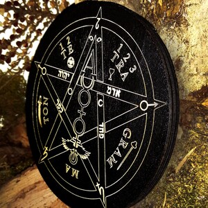 May include: A black wooden circle with a gold pentagram design. The pentagram has Hebrew text, symbols, and the words "TRA", "GRAM", "NOL", and "MA".