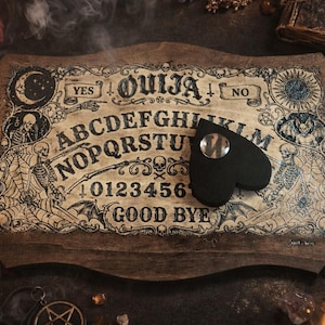 Könnte beinhalten: Ein hölzernes Ouija-Brett mit einer herzförmigen Planchette. Das Brett zeigt das Alphabet, Zahlen und die Wörter "JA" und "NEIN". Das Design umfasst Illustrationen von Skeletten, Monden und Sonnen. Das Brett ist braun mit schwarzen und weißen Details.