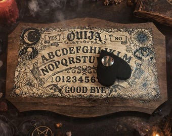 May include: A wooden Ouija board with a heart-shaped planchette. The board features the alphabet, numbers, and the words "YES" and "NO". The design includes illustrations of skeletons, moons, and suns. The board is brown with black and white details.