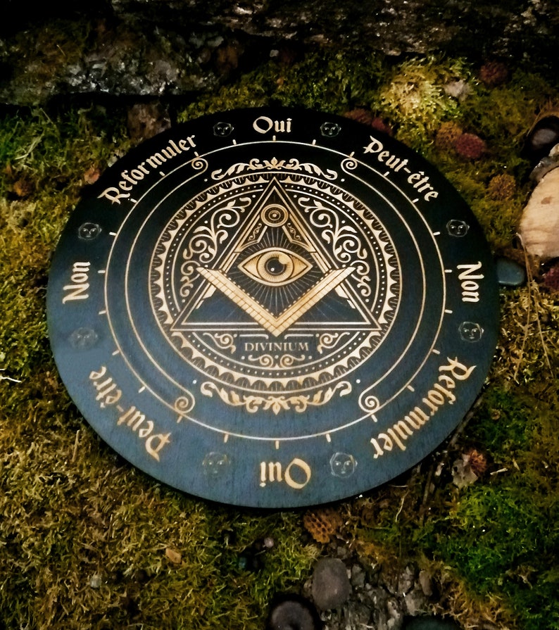 May include: A black wooden Ouija board with gold lettering and an all-seeing eye design. The board has the words "Oui", "Peut-&ecirc;tre", "Non", "Reformuler", and "Divinum" inscribed on it.