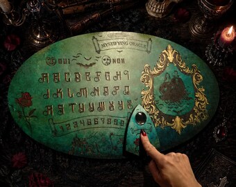 May include: An oval green spirit board titled Mystifying Oracle with gold lettering, alphabet, numbers, and a central illustration of a couple in a gold frame. A matching planchette with a clear lens rests on the board surface.