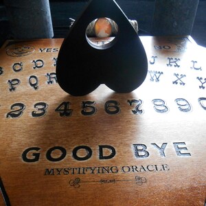 May include: A wooden Ouija board with a black heart-shaped planchette. The board features the alphabet, numbers 1-9 and 0, and the words "YES", "NO", and "GOOD BYE". The board also has the text "MYSTIFYING ORACLE" printed on it.