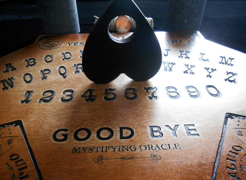 May include: A wooden Ouija board with a black heart-shaped planchette. The board features the alphabet, numbers 1-9 and 0, and the words "YES", "NO", and "GOOD BYE". The board also has the text "MYSTIFYING ORACLE" printed on it.