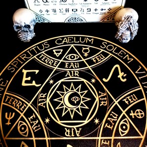 May include: A black and gold alchemical circle with symbols and text, including "SPIRITUS CAELUM SOLEM". Two small skull figurines flank the circle. A paper with alchemical symbols is in the background.