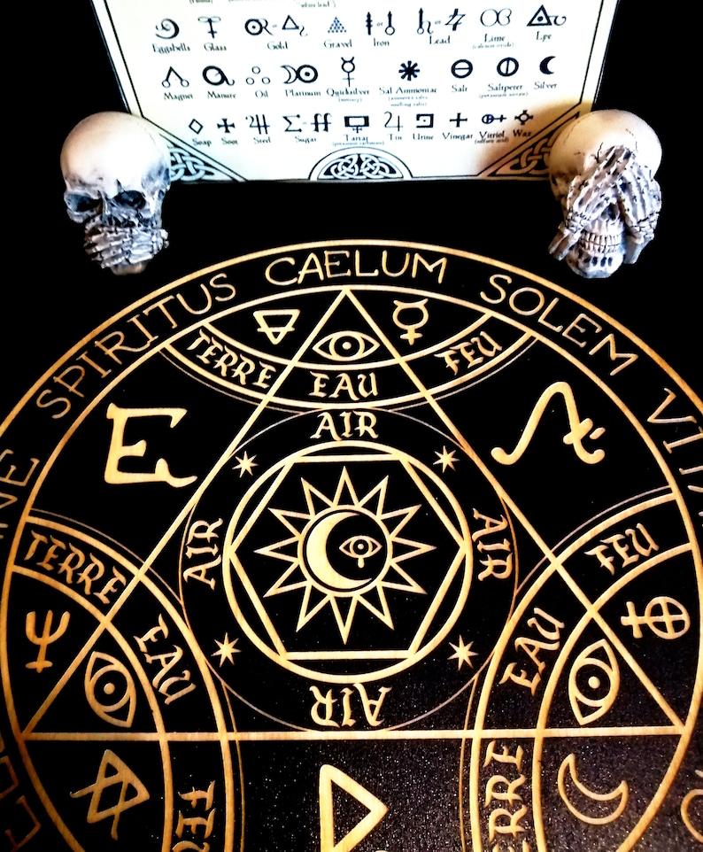 May include: A black and gold alchemical circle with symbols and text, including "SPIRITUS CAELUM SOLEM". Two small skull figurines flank the circle. A paper with alchemical symbols is in the background.