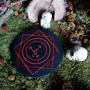 Op de afbeelding: Een ronde zwarte houten onderzetter met een rood pentagram-ontwerp. De onderzetter is omgeven door mos en kleine decoratieve schedels.