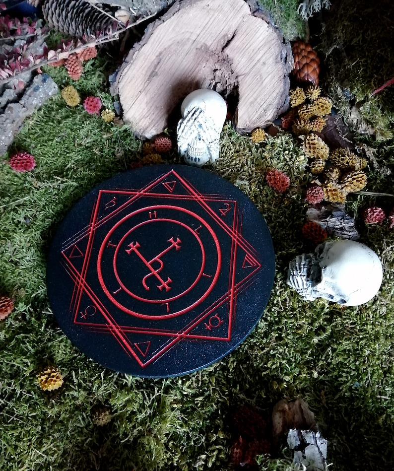 Op de afbeelding: Een ronde zwarte houten onderzetter met een rood pentagram-ontwerp. De onderzetter is omgeven door mos en kleine decoratieve schedels.