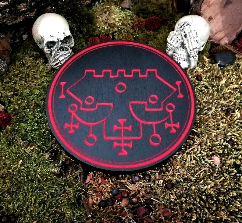 May include: A black circular wooden plaque with a red sigil design. The sigil is a complex geometric pattern with a central circle and multiple lines and symbols. The plaque is surrounded by a red border.
