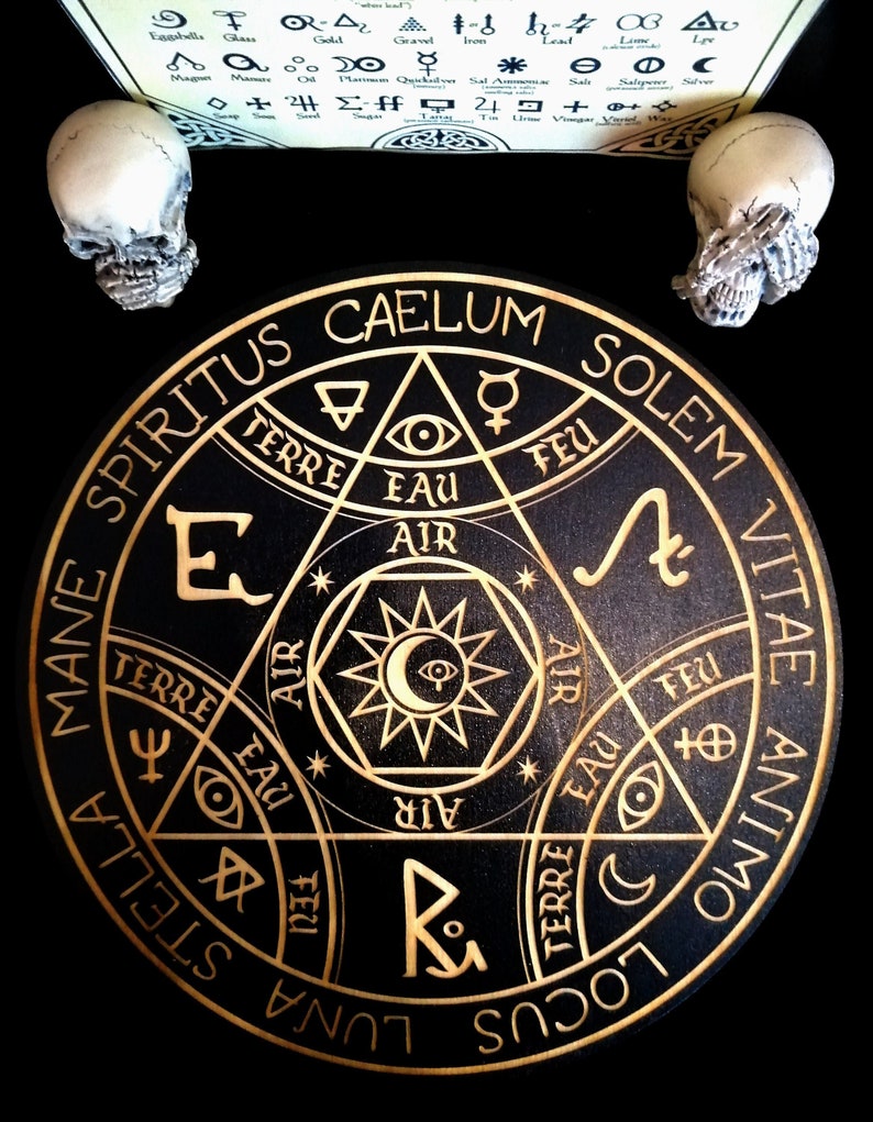 May include: A black wooden circular decorative piece with gold-colored etched symbols and text, including "CAELUM" and "SOLEMVITAE". Two small skull figurines flank the piece, with a scroll of symbols above. The overall theme is occult or esoteric.