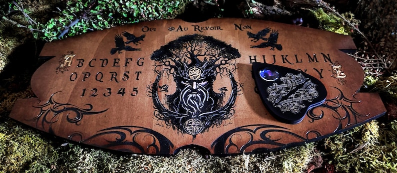 May include: A wooden Ouija board with a black and white illustration of a tree spirit. The board features the letters A-Z, numbers 1-9, and the words "Oui", "Yes", "No", "Au Revoir", and "Goodbye". A black heart-shaped planchette is included.