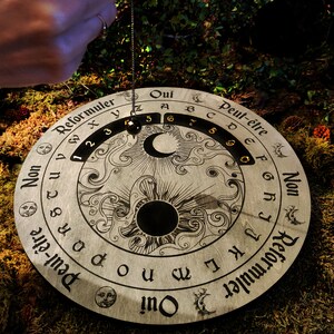 May include: A wooden pendulum board with etched celestial and numerical designs. The board features the words "Oui", "Non", and "Peut-&ecirc;tre", along with numbers and letters. A pendulum hangs above the board, ready for use. The board is circular and rests on a bed of moss.