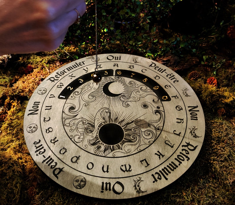 May include: A wooden pendulum board with etched celestial and numerical designs. The board features the words "Oui", "Non", and "Peut-&ecirc;tre", along with numbers and letters. A pendulum hangs above the board, ready for use. The board is circular and rests on a bed of moss.