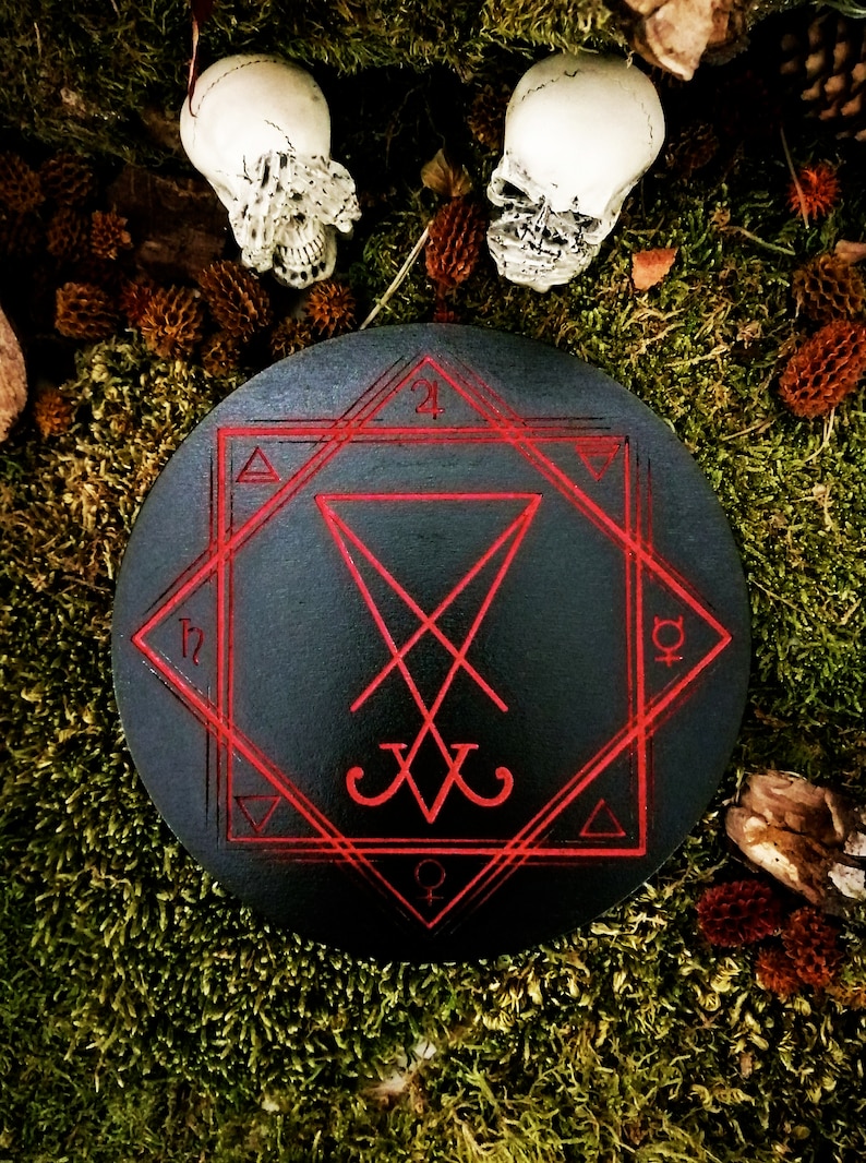 Pode incluir: Um c&iacute;rculo de madeira preto com um pentagrama vermelho e um selo inscrito nele. O c&iacute;rculo &eacute; cercado por musgo e duas caveiras brancas.