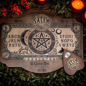 May include: A wooden Ouija board with the word "Salem" and a planchette. The board features letters, numbers, and symbols, including a crescent moon, a star, and a cat. The planchette has the word "Salem" on it. The board is surrounded by candles and greenery.