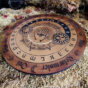 May include: A wooden Ouija board with a black and white design. The board features a hand with an eye, a sun, and a moon. The letters of the alphabet, numbers 0-9, and the words "Yes", "No", "Goodbye", and "Hello" are printed on the board.