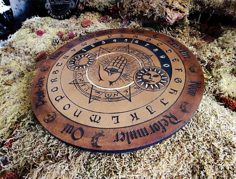 May include: A wooden Ouija board with a black and white design. The board features a hand with an eye, a sun, and a moon. The letters of the alphabet, numbers 0-9, and the words "Yes", "No", "Goodbye", and "Hello" are printed on the board.