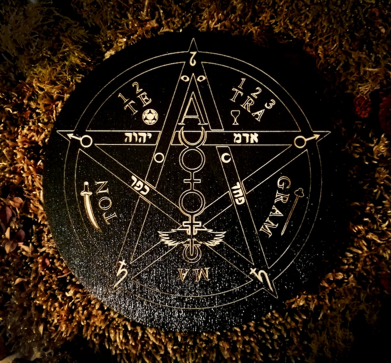 May include: A black wooden circle with a gold pentagram design. The pentagram has Hebrew letters and symbols around it. The circle also has the words "GRAM", "TRA", "TE", "NOL", and "MAM" around the outside.