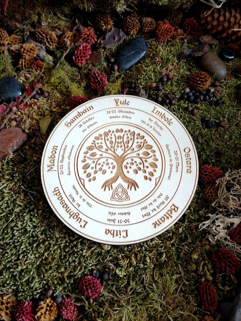 May include: A wooden circle with a tree of life design and the eight sabbats of the Wheel of the Year in French. The sabbats are Yule, Imbolc, Ostara, Beltane, Litha, Lughnasadh, Mabon, and Samhain.
