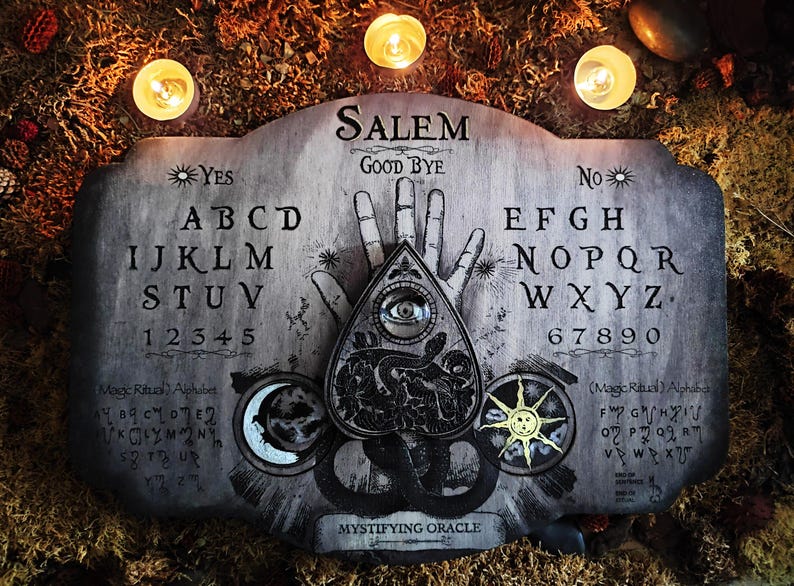 K&ouml;nnte beinhalten: Ein Ouija-Brett aus Holz mit einer schwarzen Planchette, die ein Auge und eine Schlange zeigt. Das Brett hat einen grauen Hintergrund mit schwarzen Buchstaben und Zahlen. Die W&ouml;rter "Salem" und "Mystifying Oracle" sind auf dem Brett gedruckt. Das Brett ist von drei brennenden Kerzen umgeben.