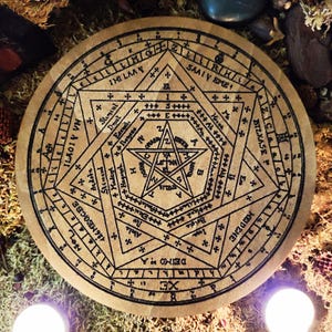 Pode incluir: Um círculo de madeira com um desenho em preto e branco de um pentagrama e outros símbolos. O círculo é rodeado por um anel de texto, incluindo as palavras "HEIDENE", "BIZKASE", "SAALY EME" e "ILAGIV VN".