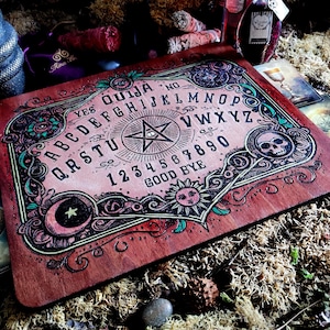May include: A wooden Ouija board with a pink and green design. The board features a pentagram, a skull, a crescent moon, and a sun. The letters of the alphabet, numbers 1-9 and 0, and the words "yes", "no", "good bye" are printed on the board.