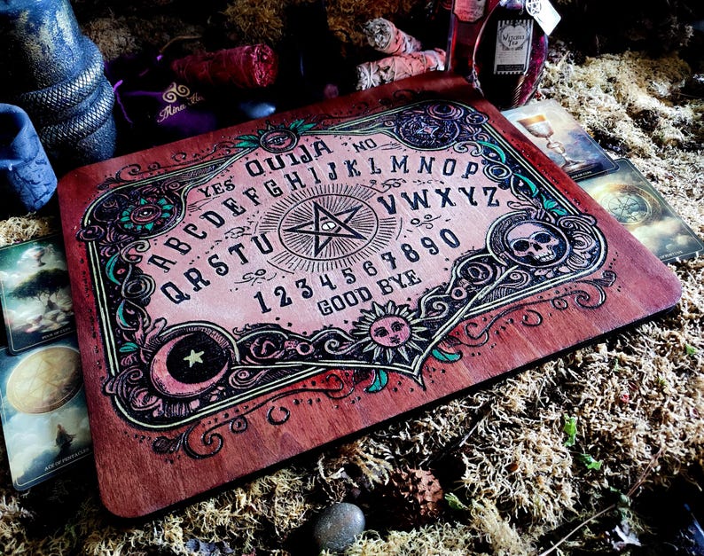 May include: A wooden Ouija board with a pink and green design. The board features a pentagram, a skull, a crescent moon, and a sun. The letters of the alphabet, numbers 1-9 and 0, and the words "yes", "no", "good bye" are printed on the board.