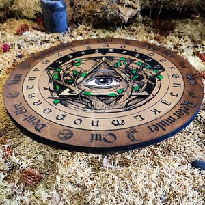 May include: A wooden Ouija board with a black and white illustration of an eye inside a triangle surrounded by branches and leaves. The board has the letters of the alphabet, numbers, and the words "Yes", "No", "Goodbye", and "Reformuler Qui" printed around the edge.