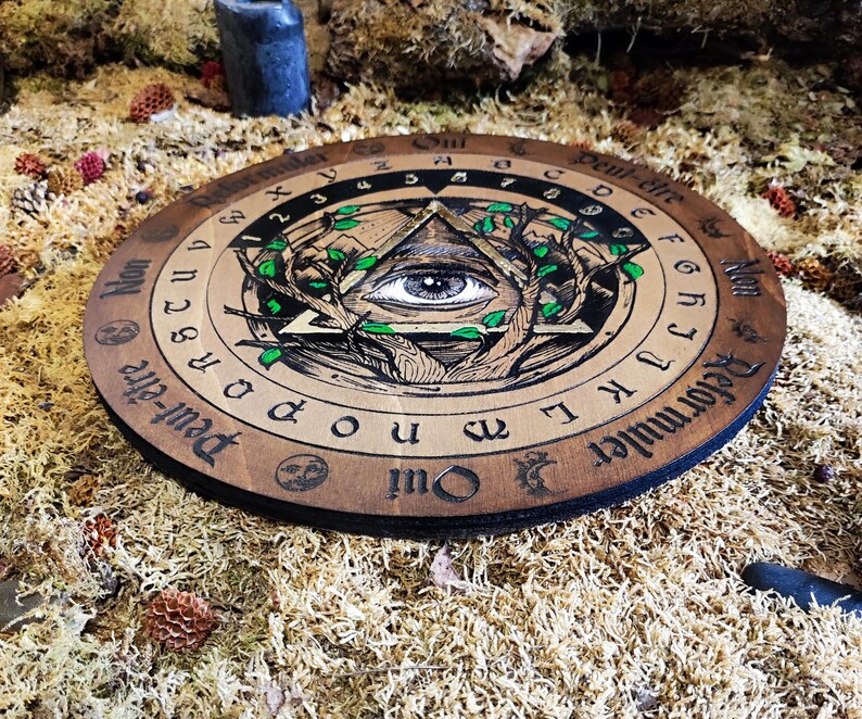 May include: A wooden Ouija board with a black and white illustration of an eye inside a triangle surrounded by branches and leaves. The board has the letters of the alphabet, numbers, and the words "Yes", "No", "Goodbye", and "Reformuler Qui" printed around the edge.