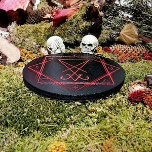Pode incluir: Um c&iacute;rculo de madeira preto com um pentagrama vermelho e um selo inscrito nele. Duas estatuetas de cr&acirc;nios brancos s&atilde;o colocadas de cada lado do c&iacute;rculo, que est&aacute; sentado em um leito de musgo verde.