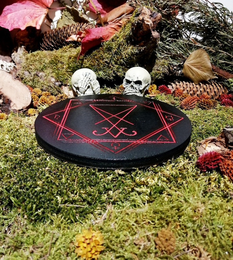 Pode incluir: Um c&iacute;rculo de madeira preto com um pentagrama vermelho e um selo inscrito nele. Duas estatuetas de cr&acirc;nios brancos s&atilde;o colocadas de cada lado do c&iacute;rculo, que est&aacute; sentado em um leito de musgo verde.