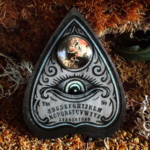 May include: A wooden planchette in a teardrop shape, featuring an eye design and the alphabet. The planchette has a dark border with a light blue accent. A clear, round lens is set in the top. The words "Yes" and "No" are on the planchette.