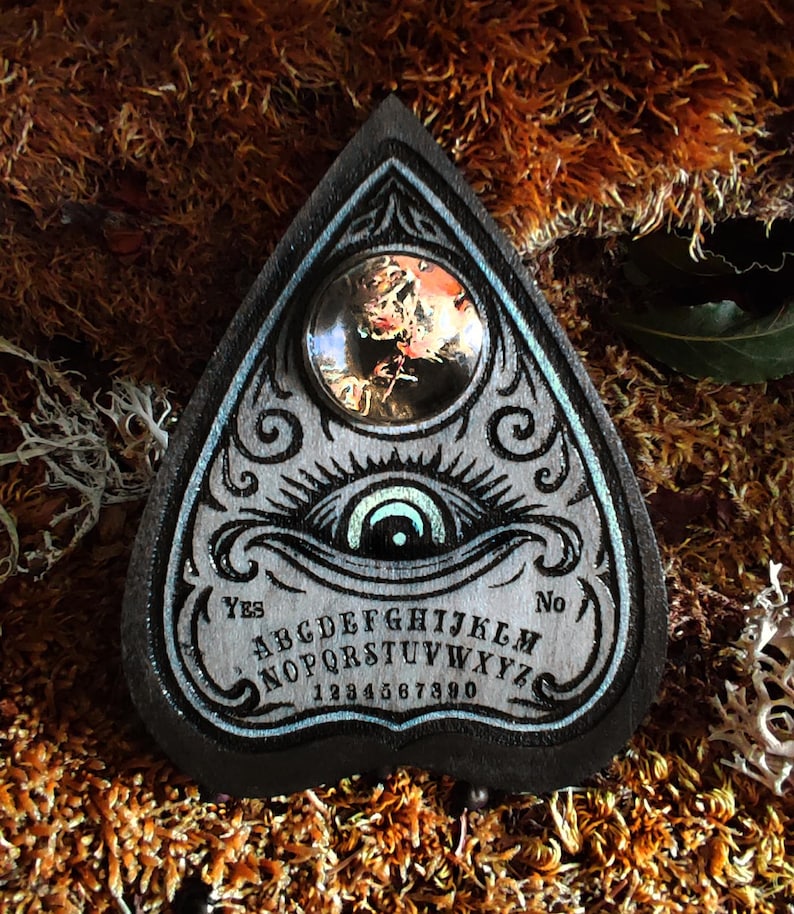 May include: A wooden planchette in a teardrop shape, featuring an eye design and the alphabet. The planchette has a dark border with a light blue accent. A clear, round lens is set in the top. The words "Yes" and "No" are on the planchette.