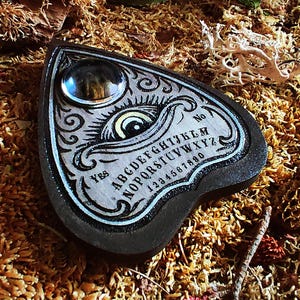 May include: A heart-shaped planchette with a decorative design. The planchette is black with a light gray center, featuring an eye illustration, the alphabet, numbers, and the words "Yes" and "No". A clear, round glass piece is at the top.