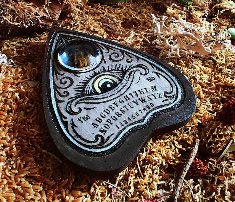 May include: A heart-shaped planchette with a decorative design. The planchette is black with a light gray center, featuring an eye illustration, the alphabet, numbers, and the words "Yes" and "No". A clear, round glass piece is at the top.