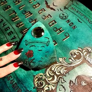 May include: A teal Ouija board with gold lettering and a planchette. The board features a decorative frame, bats, and the words "Mystifying Oracle." The planchette has a clear lens and a painted rose.