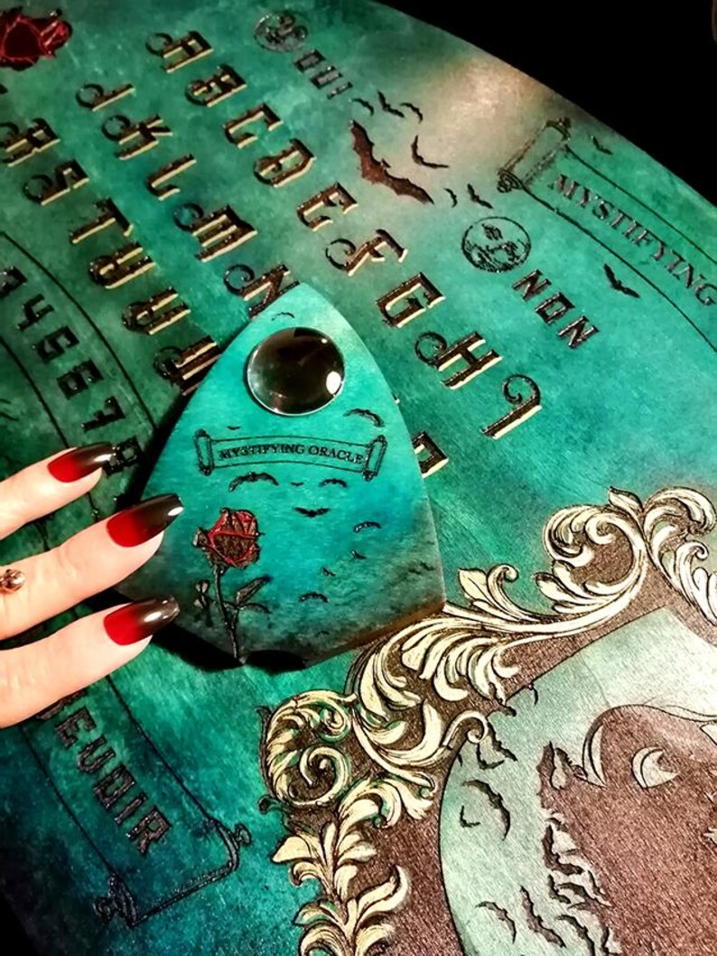 May include: A teal Ouija board with gold lettering and a planchette. The board features a decorative frame, bats, and the words "Mystifying Oracle." The planchette has a clear lens and a painted rose.