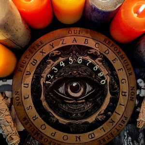 May include: A wooden Ouija board with a black and gold design featuring an eye and the letters of the alphabet, numbers 1-9 and 0, and the words "Oui", "Non", "Peut-&ecirc;tre", "Reformuler", and "Reformuler".