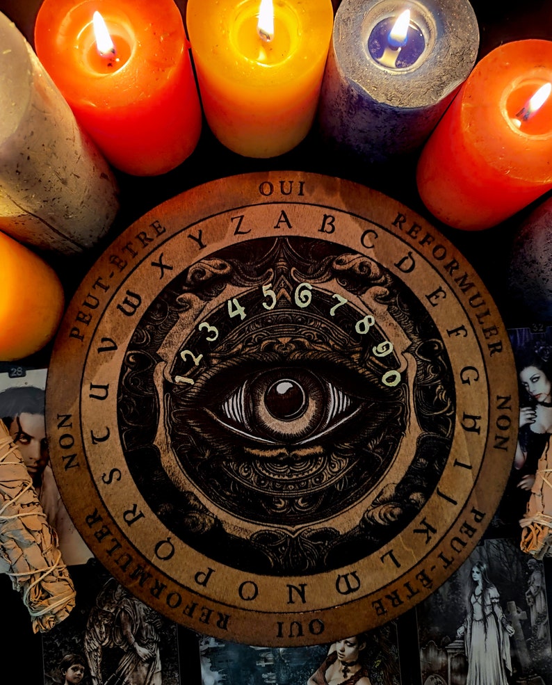 May include: A wooden Ouija board with a black and gold design featuring an eye and the letters of the alphabet, numbers 1-9 and 0, and the words "Oui", "Non", "Peut-&ecirc;tre", "Reformuler", and "Reformuler".
