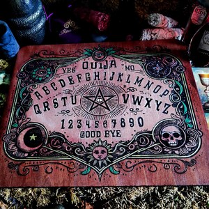 May include: A wooden Ouija board with a pink and green design. The board features a traditional alphabet, numbers, and the words "yes", "no", and "goodbye".
