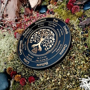 May include: A black wooden wheel with gold lettering and a tree of life design. The wheel is divided into eight sections, each representing a different Celtic holiday. The holidays are: Yule, Imbolc, Ostara, Beltane, Litha, Lughnasadh, Mabon, and Samhain.